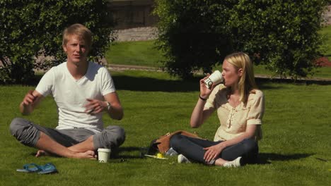 Close-up-of-two-young-blonde-students-sitting-on-grass-drinking-coffee-and-talking-as-they-enjoy-a-sunny-afternoon-outside