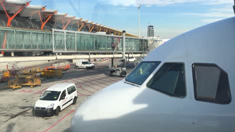 Nose-of-modern-airliner,-a-passenger-boarding-bridge-in-background,-people-boarding-an-airplane,-service-vehicles-on-tarmac,-transportation-and-travel-concept,-Madrid-Barajas-Airport