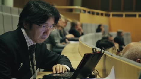Asian-man-with-glasses-looks-at-his-laptop-computer-during-a-presentation-in-a-large-modern-university-classroom-with-stadium-seating