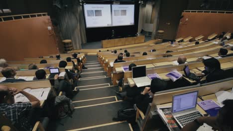 Wide-view-from-the-rear-of-a-large-modern-university-classroom-with-stadium-seating-as-meeting-attendees-listen-to-a-presentation