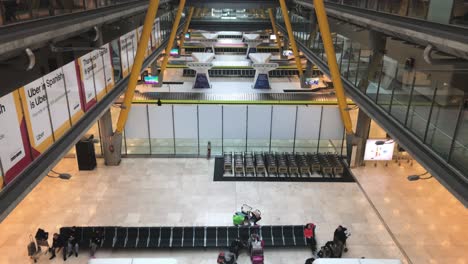 Revealing-shot-of-airport-terminal-area-with-several-baggage-claim-conveyors,-slow-tilt-up-showing-the-terminal-with-passengers-and-shops-at-the-upper-terminal-level-at-Madrid-Barajas-Airport