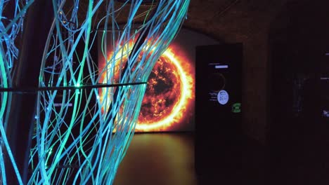 Schwenkaufnahme,-Die-Animierte-Computerbildschirme-Zeigt,-Die-Berühmte-Irische-Astronomen-Und-Ihre-Arbeit-Hinter-Der-Türkisblau-Leuchtenden-Glasfaser-Kunstskulptur-Sonne-Und-Galaxie-Auf-Großen-Bildschirmen-In-Einem-Dunklen-Raum-Zeigen