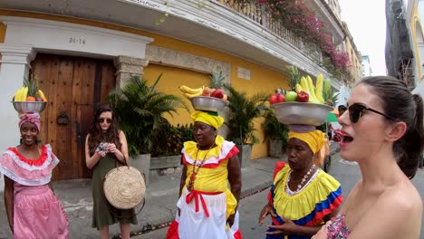 Touristen-Stehen-Und-Unterhalten-Sich-Mit-Palenqueras-In-Bunten-Kleidern,-Die-In-Ihren-Köpfen-Obstschalen-In-Der-Altstadt-Von-Cartagena-De-Indias,-Kolumbien,-Balancieren