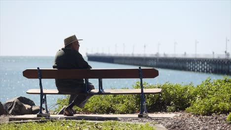 Old-man-sitting-on-bench-looking-out-to-sea-with-pier-and-blue-horizon