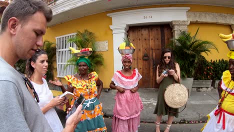 Touristen-Stehen-Und-Unterhalten-Sich-Mit-Palenqueras,-Die-Bunte-Kleider-Tragen-Und-Eine-Obstschale-über-Ihren-Köpfen-Balancieren,-In-Der-Altstadt-Von-Cartagena-De-Indias,-Kolumbien