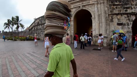Ein-Mann-Mit-Grünem-Hemd-Trägt-In-Seinem-Kopf-Dutzende-Hüte-Zum-Verkauf-In-Der-Nähe-Eines-Alten-Bogens-Und-Einer-Steinmauer,-Der-Den-Eingang-Eines-Platzes-In-Der-Altstadt-Von-Cartagena-De-Indias-Darstellt
