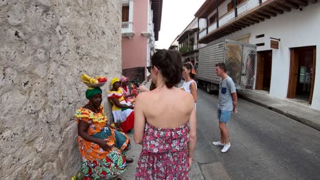 Eine-Gruppe-Touristen-Nähert-Sich-Mehreren-Palenqueras,-Die-In-Bunten-Kleidern-In-Der-Kurve-Sitzen-Und-Obstschalen-Im-Kopf-Balancieren