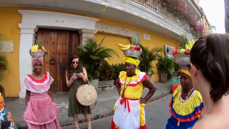 Zwei-Touristen-Stehen-Und-Sprechen-Mit-Drei-Palenqueras,-Die-Bunte-Kleider-Tragen-Und-Obstschalen-über-Ihren-Köpfen-Balancieren,-In-Der-Altstadt-Von-Cartagena-De-Indias,-Kolumbien