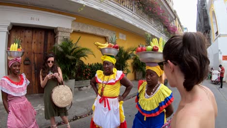 Eine-Gruppe-Bunt-Gekleideter-Palenqueras-Balanciert-Obstschalen-Auf-Ihren-Köpfen-Und-Unterhält-Sich-Dabei-Mit-Zwei-Frauen,-Die-In-Der-Altstadt-Von-Cartagena-De-Indias,-Kolumbien,-Tourismus-Betreiben