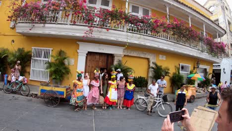 Ein-Mann-Fotografiert-Mit-Seinem-Handy-Palenqueras-Und-Weitere-Touristen-Neben-Einem-Gelben-Haus-Und-Balkonen-Mit-Blumen-In-Der-Altstadt-Von-Cartagena-De-Indias