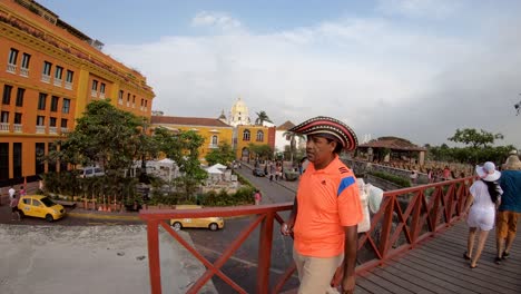 A-man-with-an-orange-shirt-and-a-hat-is-carrying-an-ice-cream-cooler-while-walking-on-a-red-bridge-of-the-old-town-of-Cartagena-de-Indias,-Colombia