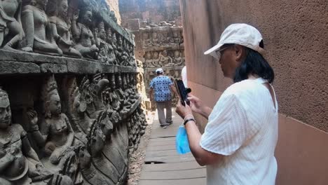 Eine-Touristin-Hält-Die-Maske-Auf-Ihrer-Hand,-Während-Sie-Mit-Ihrem-Handy-Die-Steinernen-Buddhas-Aufzeichnet,-Die-Die-Gänge-Der-Elefantenterrasse-In-Siem-Reap,-Kambodscha,-Bewachen