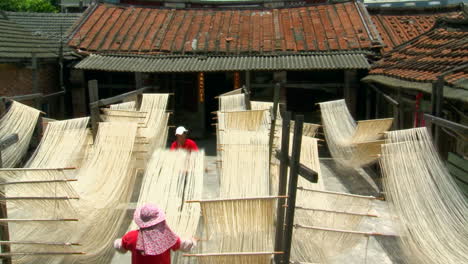 Workers-carry-strands-of-soft-fresh-noodles-into-courtyard-on-long-poles,-shake-noodles-then-hang-in-racks-to-dry-in-sunlight