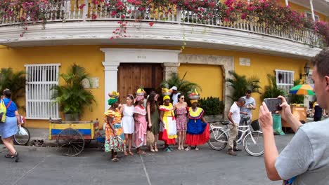 Ein-Mann-Fotografiert-Drei-Palenqueras-Und-Drei-Frauen-Neben-Einem-Gelben-Gebäude-Mit-Balkonen-Und-Blumen-In-Der-Altstadt-Von-Cartagena-De-Indias,-Kolumbien