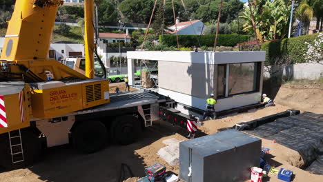 Industrial-crane-and-workers-building-modular-home-on-sunny-day,-aerial-side-view