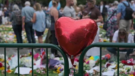 A-Red-Heart-Foil-Balloon-On-A-Fence-With-Message-Of-Condolences-To-Her-Royal-Highness,-Queen-Elizabeth-II-In-Green-Park-With-Blurred-Background-Of-People-In-London,-United-Kingdom