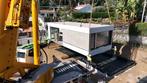Heavy-crane-and-workers-placing-down-modular-home-into-place,-aerial-descend-view