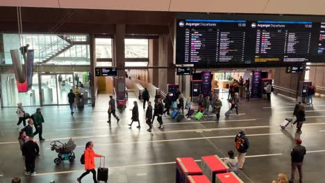 Panning-shot-of-the-inside-of-the-Oslo-Central-Station-with-lots-of-travelers-walking-with-luggage,-and-people-looking-at-the-large-screens-for-travel-information