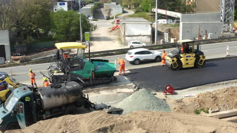 Workers-asphalting-road-with-steamroller-and-paver-next-to-car-traffic