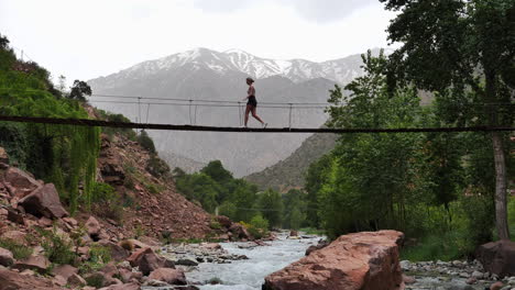 Young-adult-female-walking-across-hanging-rope-bridge-under-backdrop-of-Atlas-mountains,-Morocco