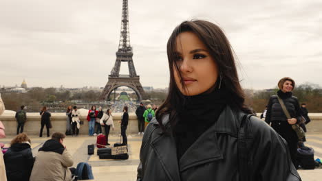 Young-brunette-girl-touching-her-hair-and-looking-at-the-camera-in-a-view-of-the-Eiffel-Tower