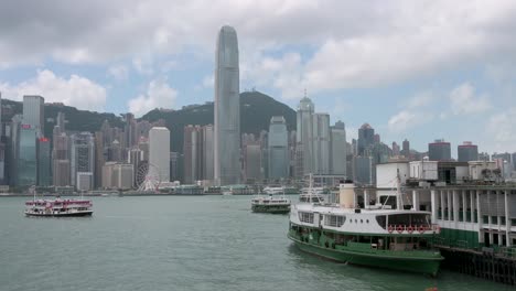 Vista-Del-Tráfico-Del-Puerto-Marítimo-De-Hong-Kong-Mientras-Los-Transbordadores-Estrella-Salen-Y-Llegan-Al-Muelle-De-La-Terminal-De-Ferry-Frente-Al-Mar-Mientras-El-Horizonte-De-Hong-Kong-Se-Ve-Al-Fondo