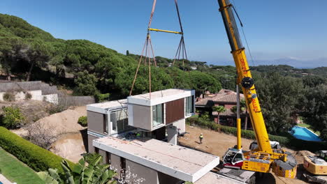 Heavy-crane-placing-modular-home-block-into-place,-aerial-fly-away-view