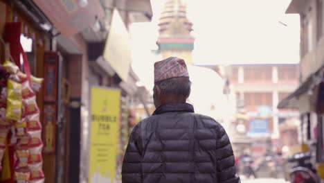 Slow-Motion-From-Behind-Of-Nepalese-Old-Man-Wearing-a-traditional-Hat-Walking-in-the-Street-With-a-Temple-in-The-Back-Kathamdu-Nepal
