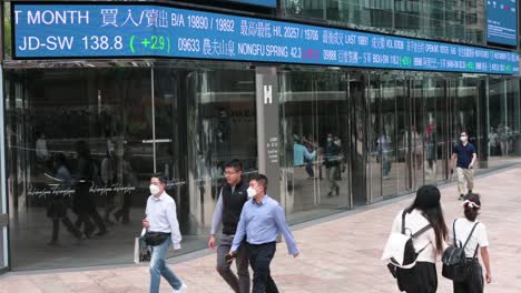 Menschen-Gehen-An-Einer-Elektronischen-Tickertafel-Und-Einem-Bildschirm-Vorbei,-Auf-Dem-Börsenzahlen-Vor-Dem-Exchange-Square-Komplex-Angezeigt-Werden,-In-Dem-Sich-Die-Hongkonger-Börse-Befindet