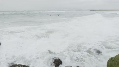 Meereswellen,-Die-Gegen-Die-Felsen-Krachen,-Und-Zwei-Surfer-Im-Hintergrund
