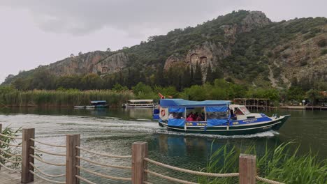 Turkish-tour-boat-speeds-on-river-under-Dalyan-Lycian-tombs-on-overcast-day