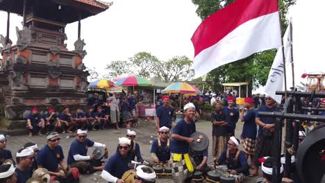 Gamelan-Musician-Hit-Percussion-Bronze-Instruments-at-Cremation-Ceremony-next-to-Indonesian-Flag-Waving-in-Hindu-Temple,-Bali-Indonesia