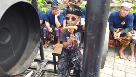Little-Kid-Plays-Gong-Musical-Gamelan-Instrument-in-Bali-Indonesia-at-Cremation-Ceremony,-Balinese-Hinduism-wearing-Glasses
