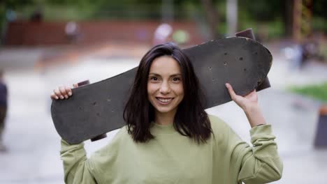 A-young-brunette-girl-stands-in-the-background-of-a-park-with-a-skateboard-and-looks-into-the-camera.-Portrait-of-a-girl