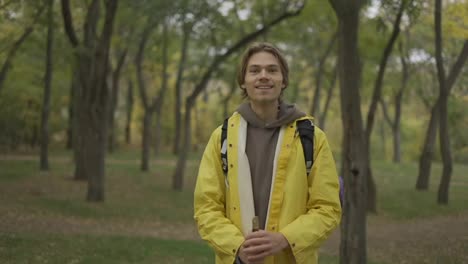 Cheerful-young-man-in-yellow-coat-with-backpack-standing-and-smiling-in-forest