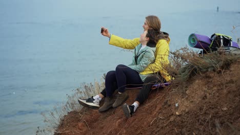 Pareja-De-Viajeros-Sentados-En-La-Cima-Del-Acantilado-Y-Posando-Tomando-Selfie-Usando-Un-Teléfono-Inteligente