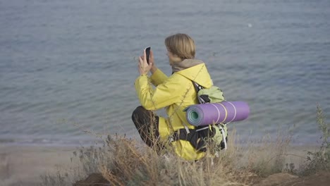 Young-man-with-backpack-takes-photo-of-the-sea-on-smartphone-while-walking-along-rocky-coast