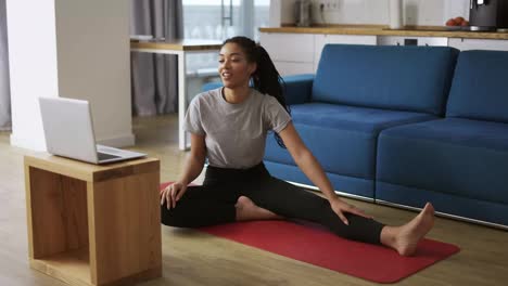 Sporty-african-american-woman-working-out,-doing-stretching-exercise-on-yoga-mat-while-watching-fitness-video