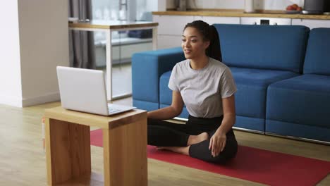 Young-african-american-woman-talking-on-video-call-on-laptop-while-sitting-on-yoga-mat