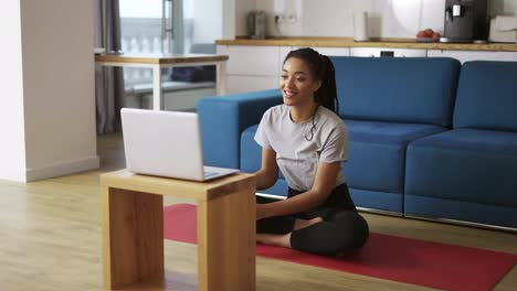 Smiling-african-american-woman-talking-on-video-call-on-laptop-while-sitting-on-yoga-mat