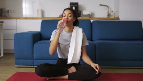 Afroamerikanische-Frau-Trinkt-Wasser-Nach-Dem-Training-Zu-Hause-Auf-Einer-Yogamatte
