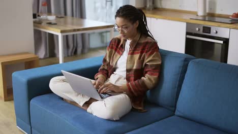 Woman-with-dreadlocks-is-working-on-new-project-and-using-laptop-on-couch-in-apartment-room