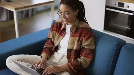 Positive-girl-with-dreadlocks-is-typing-on-laptop-on-couch,-laughing-and-smiling