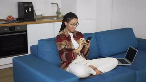 Young-woman-using-smartphone-at-home-on-a-couch-with-laptop