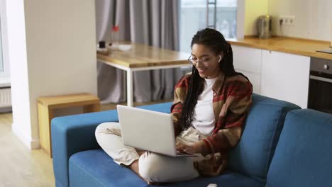Positive-girl-with-dreadlocks-sitting-on-sofa-with-laptop-on-knees