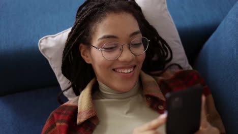 Feliz-Adolescente-Afroamericano-Relajándose-En-El-Sofá-De-Casa-Con-Un-Smartphone