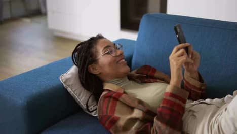 Fröhliches-Afroamerikanisches-Mädchen,-Das-Sich-Zu-Hause-Mit-Dem-Smartphone-Auf-Dem-Sofa-Entspannt