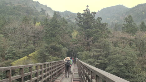 Asian-tourists-crossing-narrow-rail-track-walkway-bridge-across-lush-green-valley-in-Kyoto,-Japan