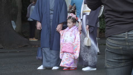 Little-Japanese-child-in-traditional-Kimono-attire-standing-still-with-two-adults-with-crowds-looking-on