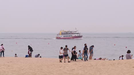 People-on-Haeundae-Beach,-ferries-passing-in-the-background
